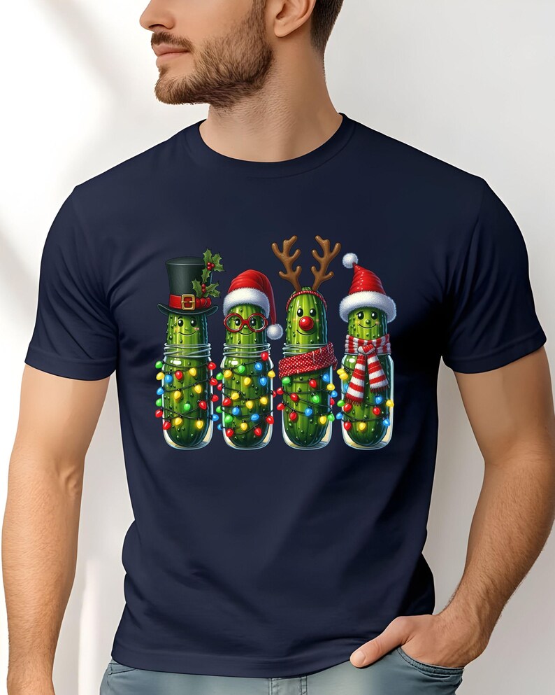 K&ouml;nnte beinhalten: Marineblaues T-Shirt mit einem weihnachtlichen Design. Die Grafik zeigt vier Gew&uuml;rzgurken in Gl&auml;sern, die jeweils mit festlichen Accessoires geschm&uuml;ckt sind: Zylinder, Weihnachtsm&uuml;tze, Rentiergeweih und Schal. Sie sind auch mit bunten Lichterketten verziert.