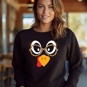 May include: Black sweatshirt featuring a turkey face design. The turkey face includes white eyes, leopard print glasses, and a yellow beak with a red wattle. The sweatshirt is a festive apparel item for Thanksgiving.