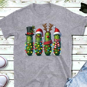 K&ouml;nnte beinhalten: Graues T-Shirt mit einem festlichen Motiv. Das Design zeigt vier Gew&uuml;rzgurken in Gl&auml;sern, die mit Weihnachtsaccessoires wie H&uuml;ten, Rentiergeweihen und Schals geschm&uuml;ckt sind. Die Gew&uuml;rzgurken sind auch mit Lichterketten umwickelt.