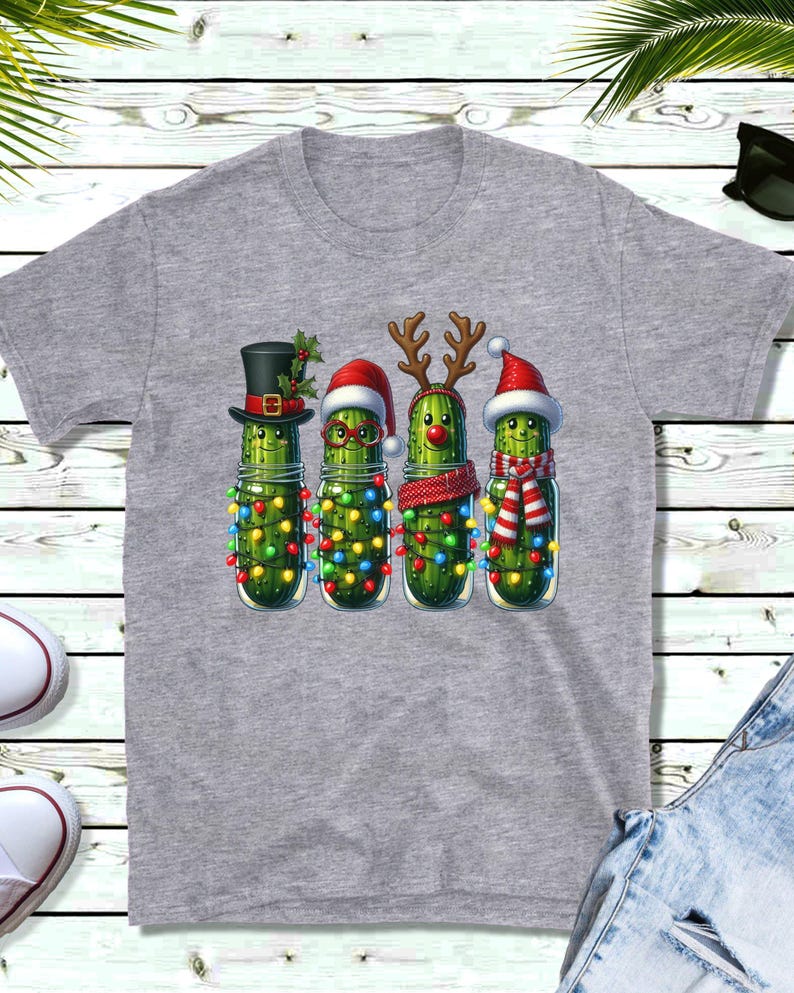 K&ouml;nnte beinhalten: Graues T-Shirt mit einem festlichen Motiv. Das Design zeigt vier Gew&uuml;rzgurken in Gl&auml;sern, die mit Weihnachtsaccessoires wie H&uuml;ten, Rentiergeweihen und Schals geschm&uuml;ckt sind. Die Gew&uuml;rzgurken sind auch mit Lichterketten umwickelt.