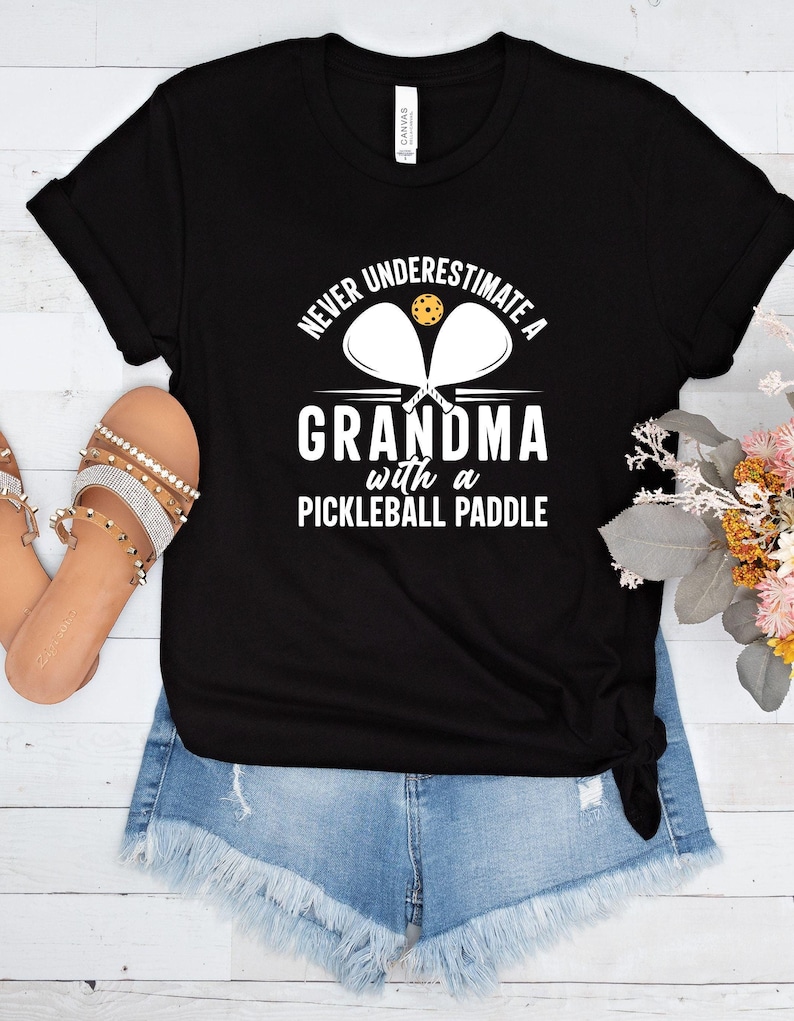 Op de afbeelding: Zwart T-shirt met witte tekst die luidt "Never underestimate a grandma with a pickleball paddle". De tekst wordt omringd door een cirkel met twee pickleball-rackets en een pickleball.