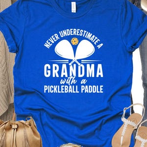 Op de afbeelding: Een koninklijk blauw T-shirt met witte tekst die "Never underestimate a grandma with a pickleball paddle" luidt. De tekst wordt vergezeld van een afbeelding van twee pickleball-rackets en een bal.