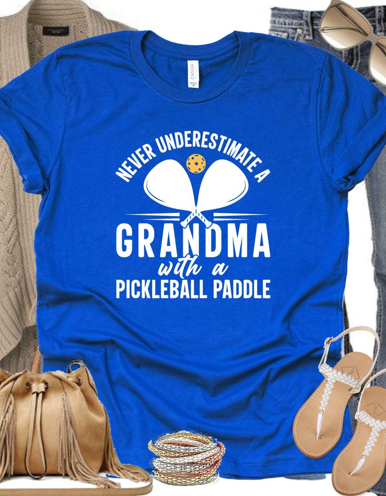Op de afbeelding: Een koninklijk blauw T-shirt met witte tekst die "Never underestimate a grandma with a pickleball paddle" luidt. De tekst wordt vergezeld van een afbeelding van twee pickleball-rackets en een bal.