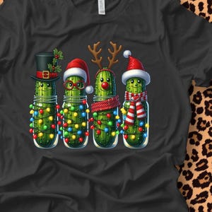 K&ouml;nnte beinhalten: Schwarzes T-Shirt mit vier Cartoon-Gurken in Gl&auml;sern, die jeweils f&uuml;r Weihnachten dekoriert sind. Eine tr&auml;gt einen Zylinder, eine andere eine Brille und einen Weihnachtsmannhut, die dritte ein Rentiergeweih und die letzte einen Weihnachtsmannhut und einen Schal. Jedes Glas ist mit bunten Weihnachtslichtern geschm&uuml;ckt.