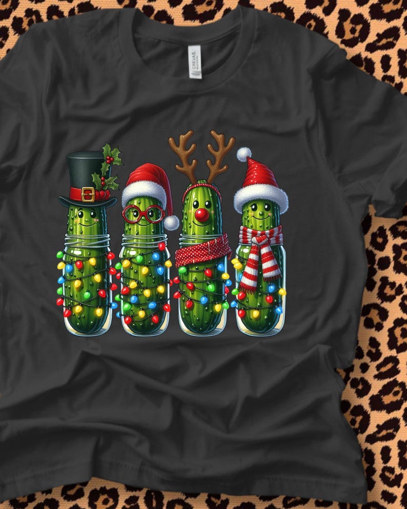 K&ouml;nnte beinhalten: Schwarzes T-Shirt mit vier Cartoon-Gurken in Gl&auml;sern, die jeweils f&uuml;r Weihnachten dekoriert sind. Eine tr&auml;gt einen Zylinder, eine andere eine Brille und einen Weihnachtsmannhut, die dritte ein Rentiergeweih und die letzte einen Weihnachtsmannhut und einen Schal. Jedes Glas ist mit bunten Weihnachtslichtern geschm&uuml;ckt.
