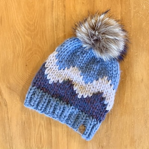 Puede incluir: Un gorro de punto azul, blanco y morado con un pompón de piel sintética. El gorro presenta un patrón en zigzag y una pequeña etiqueta de cuero. Este gorro de invierno es perfecto para el frío.