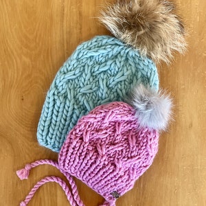 Puede incluir: Dos gorros de punto con pompones. El gorro de arriba es azul claro con un pompón marrón. El gorro de abajo es rosa con un pompón gris y tiene una larga corbata.