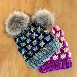 Puede incluir: Dos gorros de punto con pompones de piel sintética. El gorro de arriba es de color morado y rosa con un patrón blanco y rosa. El gorro de abajo es de color azul, verde y morado con un patrón geométrico.