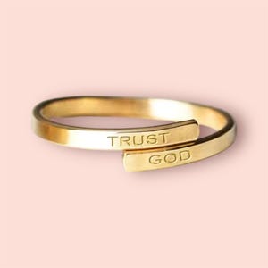 May include: A gold-toned ring with the words "TRUST GOD" engraved on it. The ring is designed to be worn on the finger with the words visible.