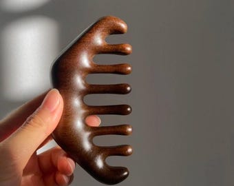 May include: A dark brown wooden hair comb with five teeth. The comb is shaped like a crescent moon and has a smooth, polished finish.