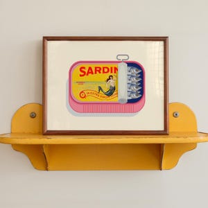 May include: A framed print of a sardine can with a pink base and a yellow label. The label reads "SARDIN" and features a mermaid illustration. The can is open, revealing sardines. The print is displayed on a yellow shelf.