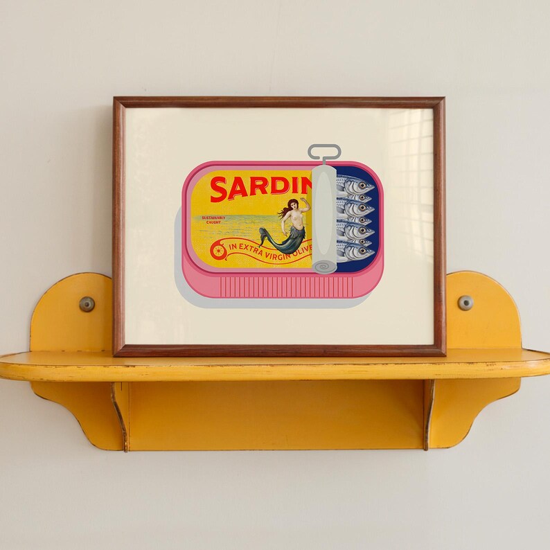 May include: A framed print of a sardine can with a pink base and a yellow label. The label reads "SARDIN" and features a mermaid illustration. The can is open, revealing sardines. The print is displayed on a yellow shelf.