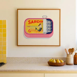 May include: A framed print of a sardine can with a pink base and a yellow label. The label reads "SARDIN" and features a mermaid illustration. The can contains several sardines. The print is displayed in a kitchen setting.