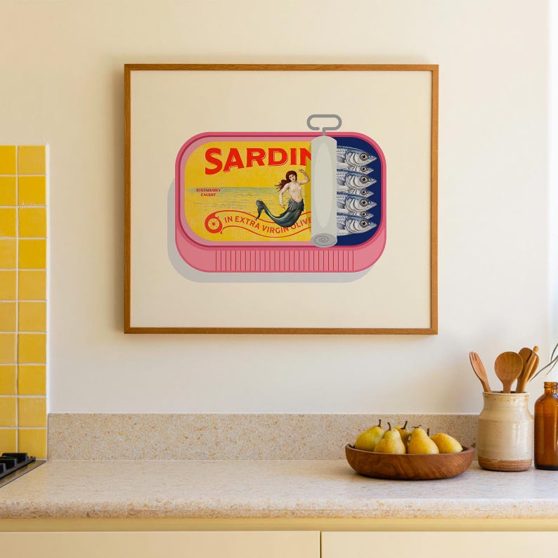 May include: A framed print of a sardine can with a pink base and a yellow label. The label reads "SARDIN" and features a mermaid illustration. The can contains several sardines. The print is displayed in a kitchen setting.
