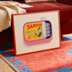 May include: A framed art print featuring a pink sardine can with the word "SARDIN" in red. The can's label depicts a mermaid and the words "IN EXTRA VIRGIN OLIVE". The can contains several fish. The frame is red.