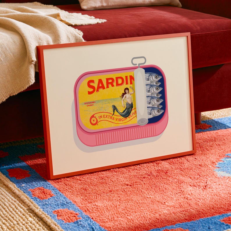 May include: A framed art print featuring a pink sardine can with the word "SARDIN" in red. The can's label depicts a mermaid and the words "IN EXTRA VIRGIN OLIVE". The can contains several fish. The frame is red.