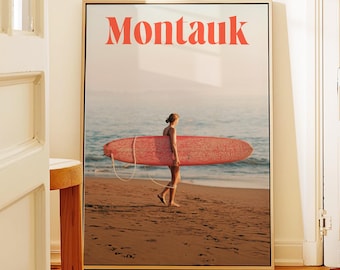 May include: A framed wall art print featuring the word Montauk in bold red letters at the top. The image shows a person walking along a sandy beach carrying a long, textured red surfboard toward the ocean waves under a soft, hazy sky.
