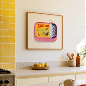 May include: A framed art print featuring a pink sardine can design with the word "Sardin" in yellow. The can is open, revealing sardines and a mermaid illustration. The print is in a wooden frame.