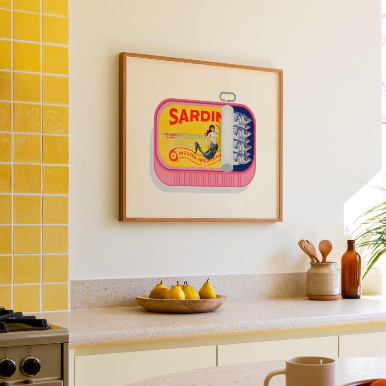 May include: A framed art print featuring a pink sardine can design with the word "Sardin" in yellow. The can is open, revealing sardines and a mermaid illustration. The print is in a wooden frame.