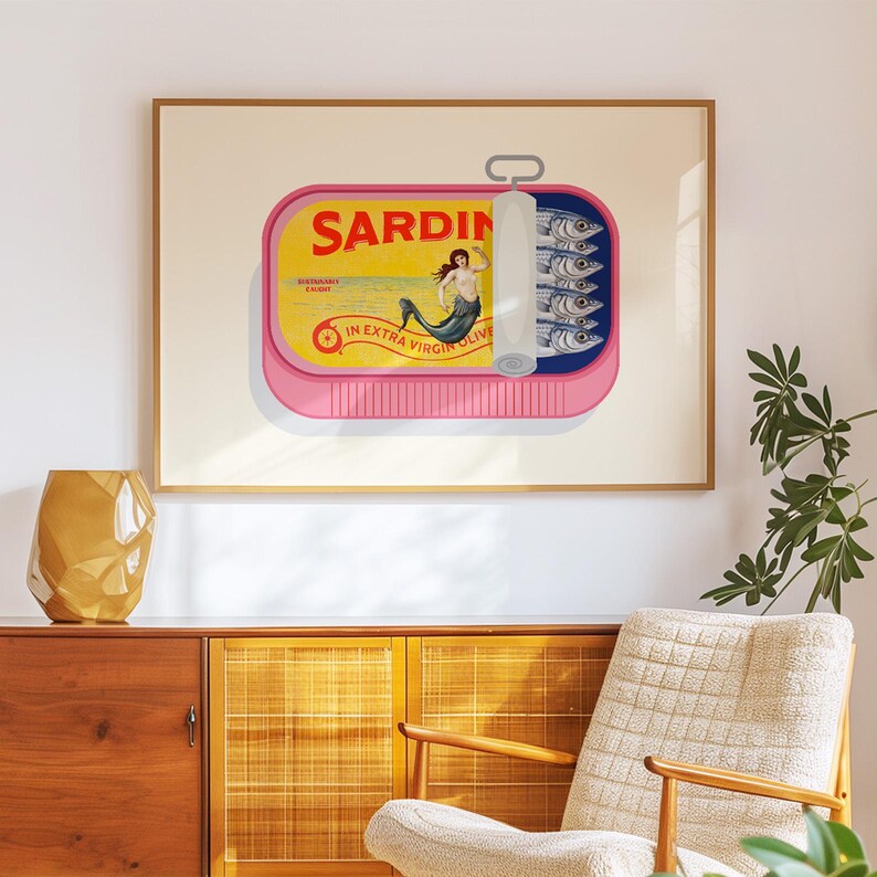 May include: A framed art print featuring a pink sardine can with a yellow label and the word "Sardines". The can is open, revealing sardines and a mermaid illustration. The print is displayed on a wall above a wooden cabinet.