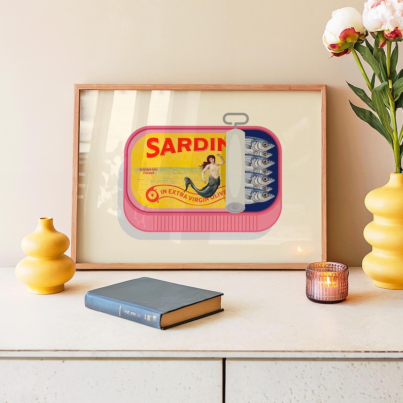 May include: A framed print featuring a pink sardine can illustration. The can is open, revealing sardines. The label reads "SARDIN" with a mermaid graphic. The print is in a wooden frame, with a yellow vase and a book in the foreground.