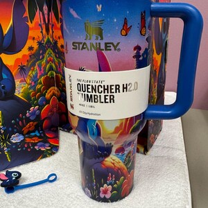 Może przedstawiać: Kubek Stanley Quencher H2.0 o pojemności 1,18 L z żywą tropikalną sceną, motylami i grafiką postaci. Kubek z niebieskim uchwytem ma złoty brzeg, ciemnoniebieską pokrywkę oraz papierową opaskę z napisem The Flowstate Quencher H2.0 Tumbler 40 oz 1.18 L.