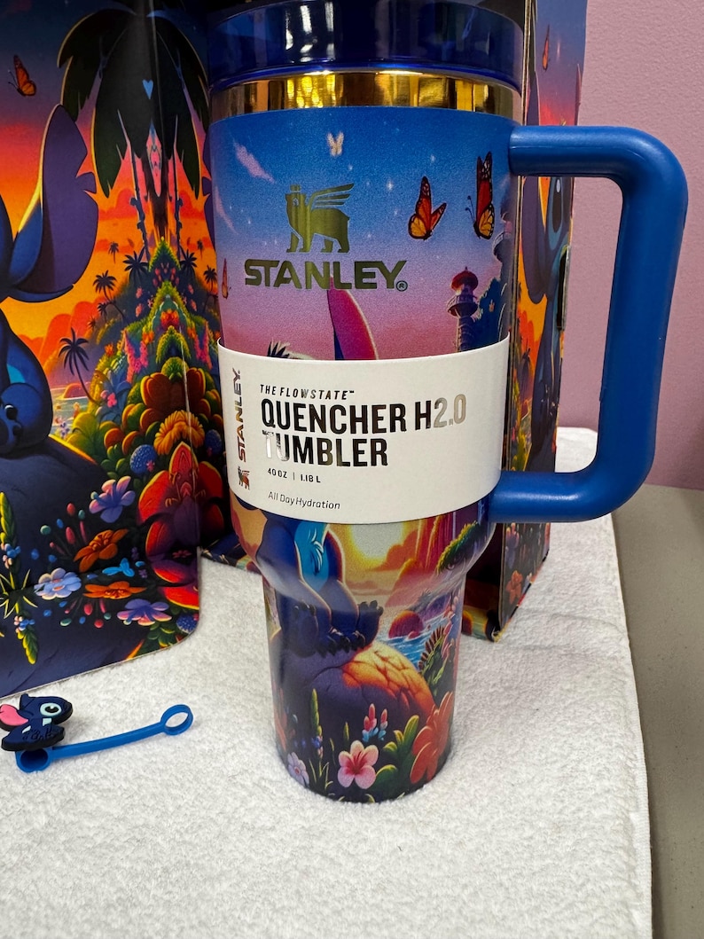 Może przedstawiać: Kubek Stanley Quencher H2.0 o pojemności 1,18 L z żywą tropikalną sceną, motylami i grafiką postaci. Kubek z niebieskim uchwytem ma złoty brzeg, ciemnoniebieską pokrywkę oraz papierową opaskę z napisem The Flowstate Quencher H2.0 Tumbler 40 oz 1.18 L.