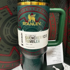 May include: A dark green Stanley Flowstate Quencher H2.0 tumbler with a 40 oz capacity. The tumbler features a gold-colored logo of a winged bear and intricate red and green geometric patterns. It includes a matching green handle and lid.