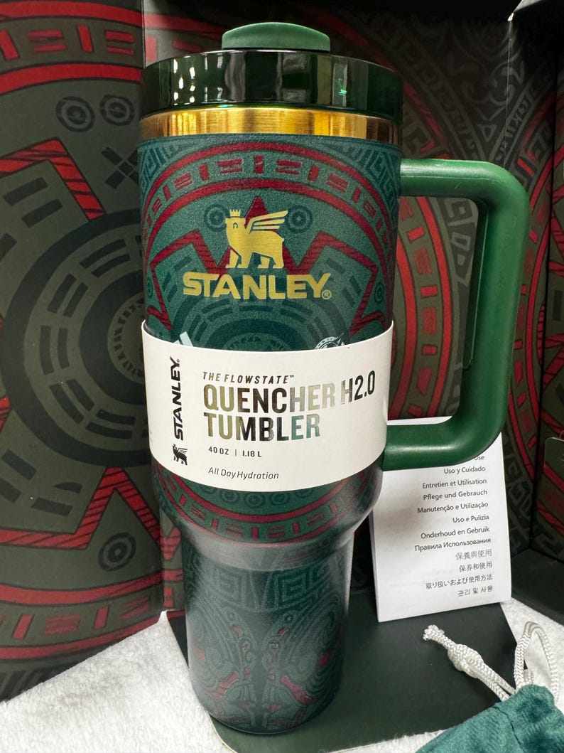May include: A dark green Stanley Flowstate Quencher H2.0 tumbler with a 40 oz capacity. The tumbler features a gold-colored logo of a winged bear and intricate red and green geometric patterns. It includes a matching green handle and lid.