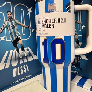 Może przedstawiać: Biało-niebieski kubek Stanley Flowstate Quencher H2.0 w paski o pojemności 1,18 L. Kubek posiada duży niebieski numer 10, złoty brzeg i biały uchwyt. Jest prezentowany na tle opakowania przedstawiającego Lionela Messiego.
