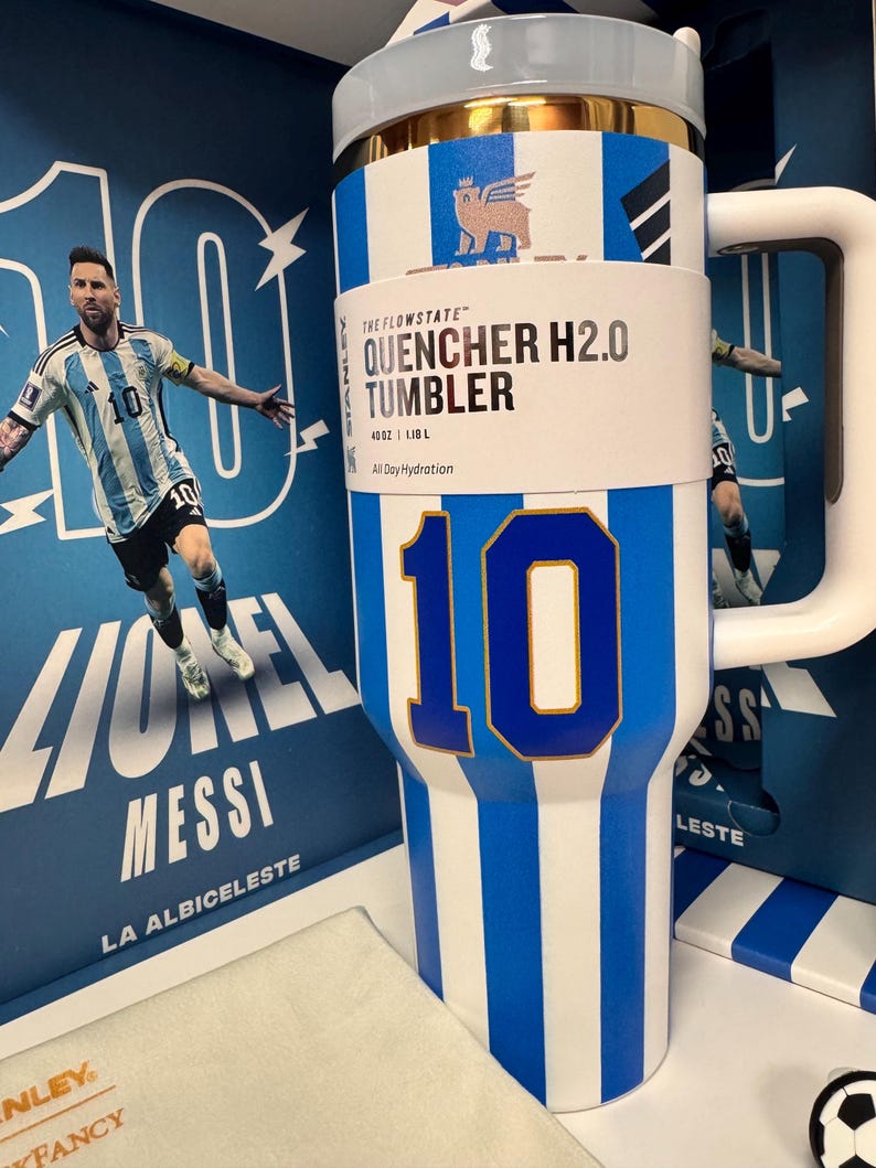Może przedstawiać: Biało-niebieski kubek Stanley Flowstate Quencher H2.0 w paski o pojemności 1,18 L. Kubek posiada duży niebieski numer 10, złoty brzeg i biały uchwyt. Jest prezentowany na tle opakowania przedstawiającego Lionela Messiego.