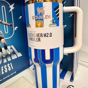 Może przedstawiać: Biało-niebieski kubek Stanley Quencher H2.0 w paski ze złotą krawędzią, ozdobiony numerem 10, logo Stanley i grafikami piłkarskimi. Kubek posiada biały uchwyt, przezroczystą pokrywkę oraz nakładkę na słomkę w kształcie piłki nożnej.