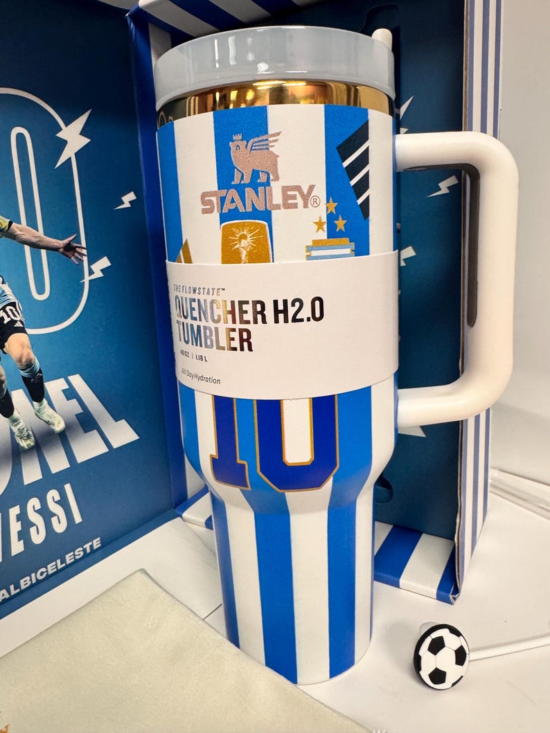 Może przedstawiać: Biało-niebieski kubek Stanley Quencher H2.0 w paski ze złotą krawędzią, ozdobiony numerem 10, logo Stanley i grafikami piłkarskimi. Kubek posiada biały uchwyt, przezroczystą pokrywkę oraz nakładkę na słomkę w kształcie piłki nożnej.