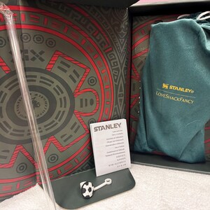 May include: A Stanley LoveShackFancy branded dark green fabric pouch, a clear plastic straw, and a small white and black soccer ball charm inside a decorative box with red and grey geometric patterns. A white Stanley care instruction card is included.