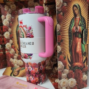 May include: A white Stanley Quencher H2.0 tumbler featuring a pink handle and a floral pattern with an image of the Virgin of Guadalupe. The tumbler is displayed in a decorative box with matching rose imagery and includes a small pink straw topper.