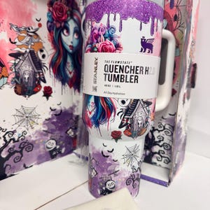May include: A 40 oz Stanley The Flowstate Quencher H2.0 tumbler featuring a white base with purple glitter drip accents and a graphic print of characters, roses, and spiderwebs. The tumbler includes a white handle, a clear lid, and a white straw.