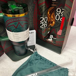 May include: A dark green insulated tumbler with a gold rim and a green lid, featuring a patterned design. It is displayed with a matching green drawstring pouch, a clear straw, and a decorative box marked with FIFA World Cup 2026 branding.