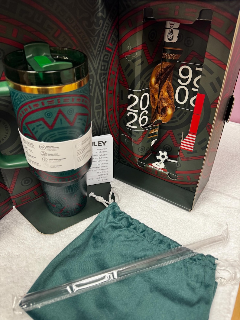 May include: A dark green insulated tumbler with a gold rim and a green lid, featuring a patterned design. It is displayed with a matching green drawstring pouch, a clear straw, and a decorative box marked with FIFA World Cup 2026 branding.