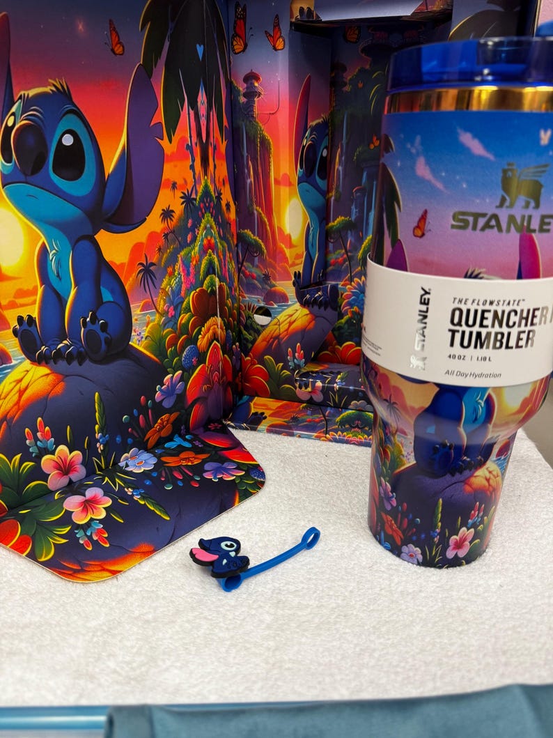 Może przedstawiać: Kubek Stanley Flowstate Quencher o pojemności 1,18 litra z kolorową tropikalną sceną przedstawiającą Stitcha, palmy i kwiaty. Kubek zawiera niebieską nakładkę na słomkę w kształcie Stitcha i jest prezentowany na tle pasującego opakowania.