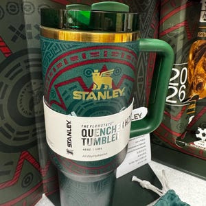May include: A 40 oz Stanley Flowstate Quencher tumbler in dark green with a gold rim and a geometric red and green pattern. The tumbler features a gold Stanley logo with a winged creature, a green handle, and a dark green lid. A white paper band displays the text The Flowstate Quencher Tumbler 40 oz 1.18 L All Day Hydration.
