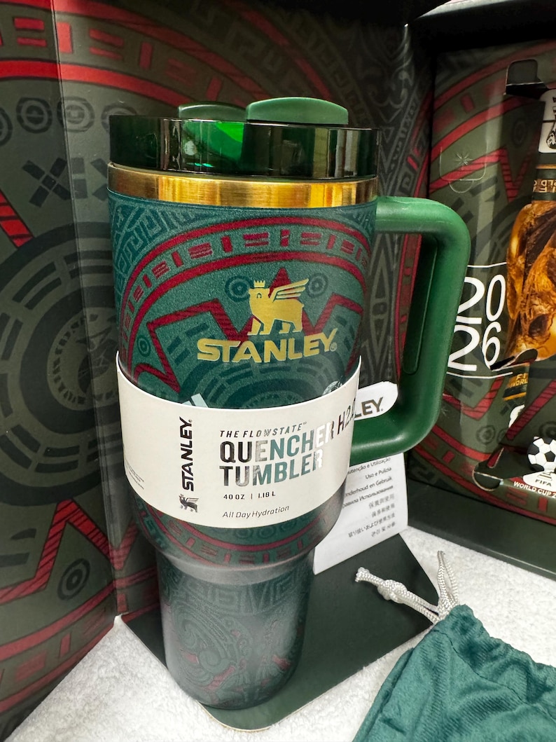 May include: A 40 oz Stanley Flowstate Quencher tumbler in dark green with a gold rim and a geometric red and green pattern. The tumbler features a gold Stanley logo with a winged creature, a green handle, and a dark green lid. A white paper band displays the text The Flowstate Quencher Tumbler 40 oz 1.18 L All Day Hydration.
