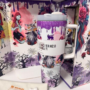 May include: A white Stanley Quencher H2.0 tumbler featuring a purple glitter drip design at the top and a Nightmare Before Christmas graphic print. The tumbler includes a white handle, a lid, and a white dust bag with Stanley LoveShackFancy text.