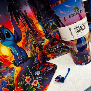 Może przedstawiać: Kubek Stanley Flowstate Quencher H2.0 o pojemności 1,18 litra, ozdobiony żywą tropikalną sceną ze Stichem, kolorowymi kwiatami i motylami. Kubek posiada niebieską pokrywkę, uchwyt oraz mały pasujący dodatek w formie zawieszki ze Stichem.