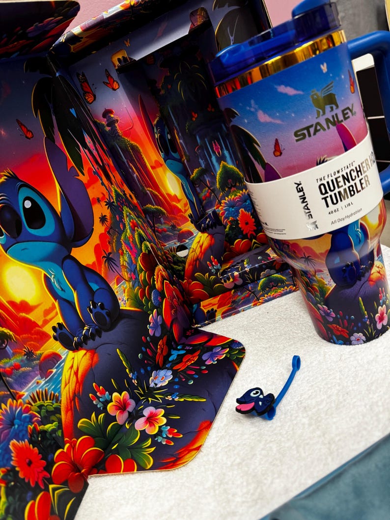 Może przedstawiać: Kubek Stanley Flowstate Quencher H2.0 o pojemności 1,18 litra, ozdobiony żywą tropikalną sceną ze Stichem, kolorowymi kwiatami i motylami. Kubek posiada niebieską pokrywkę, uchwyt oraz mały pasujący dodatek w formie zawieszki ze Stichem.