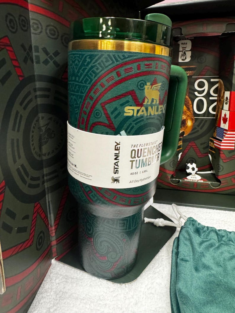 May include: A dark green Stanley Flowstate Quencher tumbler with a 40 oz capacity. The cup features a gold rim, a green handle, and a dark green lid. A white paper band wraps around the center with the text Stanley, The Flowstate Quencher, 40 oz, 1.18 L.