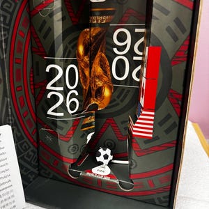 May include: A FIFA World Cup 2026 commemorative display box featuring a gold-colored trophy graphic, the text 2026, and a small black and white soccer ball model. The packaging includes red, black, and grey geometric patterns and national flag motifs.