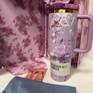 May include: A light purple Stanley Quencher H2.0 tumbler featuring a floral pattern, a gold rim, and a matching handle. The tumbler is displayed in a floral box with a blue pouch and a small card. The text on the tumbler reads Stanley, LoveShackFancy, and Quencher H2.0.