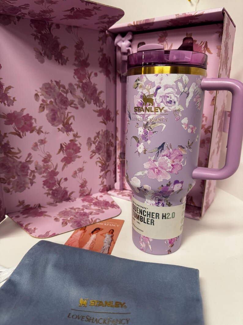 May include: A light purple Stanley Quencher H2.0 tumbler featuring a floral pattern, a gold rim, and a matching handle. The tumbler is displayed in a floral box with a blue pouch and a small card. The text on the tumbler reads Stanley, LoveShackFancy, and Quencher H2.0.