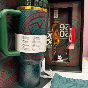 May include: A dark green stainless steel tumbler with a handle and a gold rim, featuring red and green geometric patterns. A white product information band wraps around the center. The tumbler is displayed in a box marked with 2026 and a soccer ball.