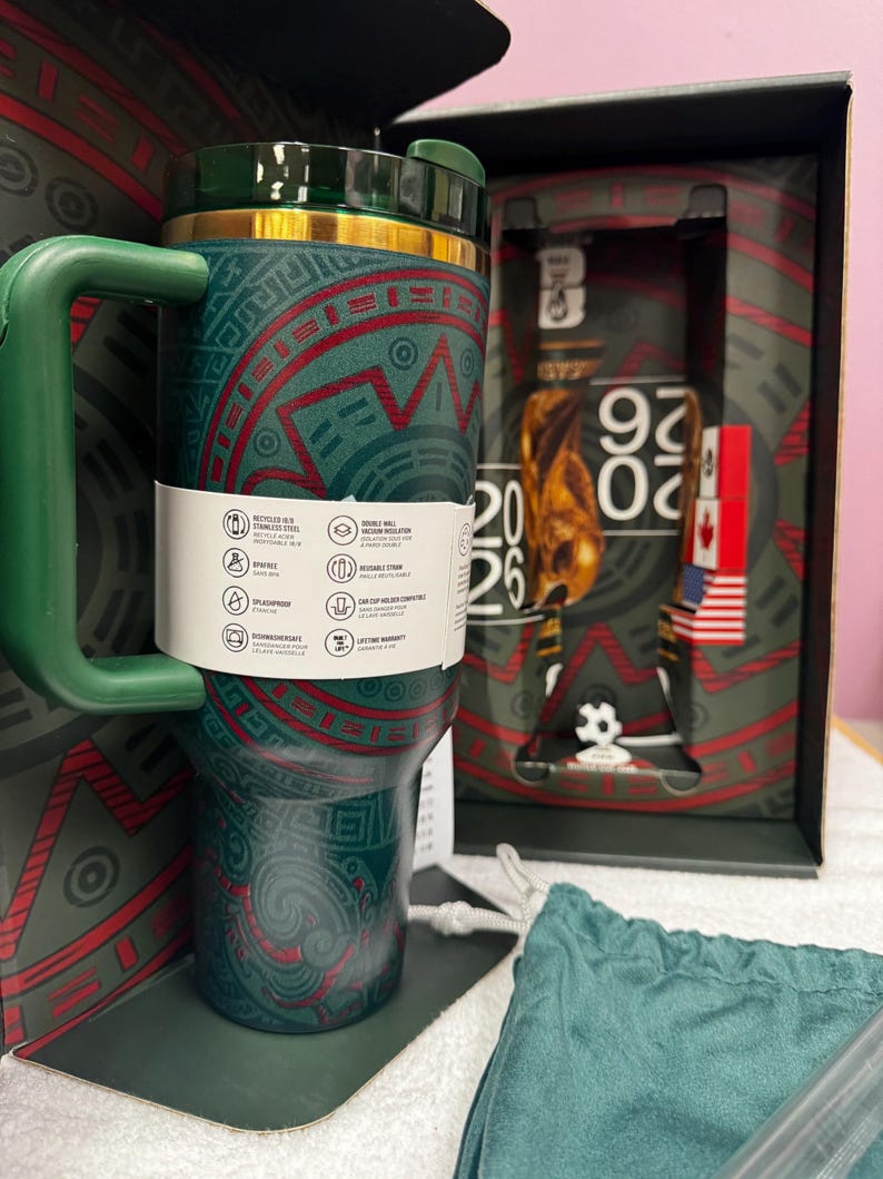 May include: A dark green stainless steel tumbler with a handle and a gold rim, featuring red and green geometric patterns. A white product information band wraps around the center. The tumbler is displayed in a box marked with 2026 and a soccer ball.